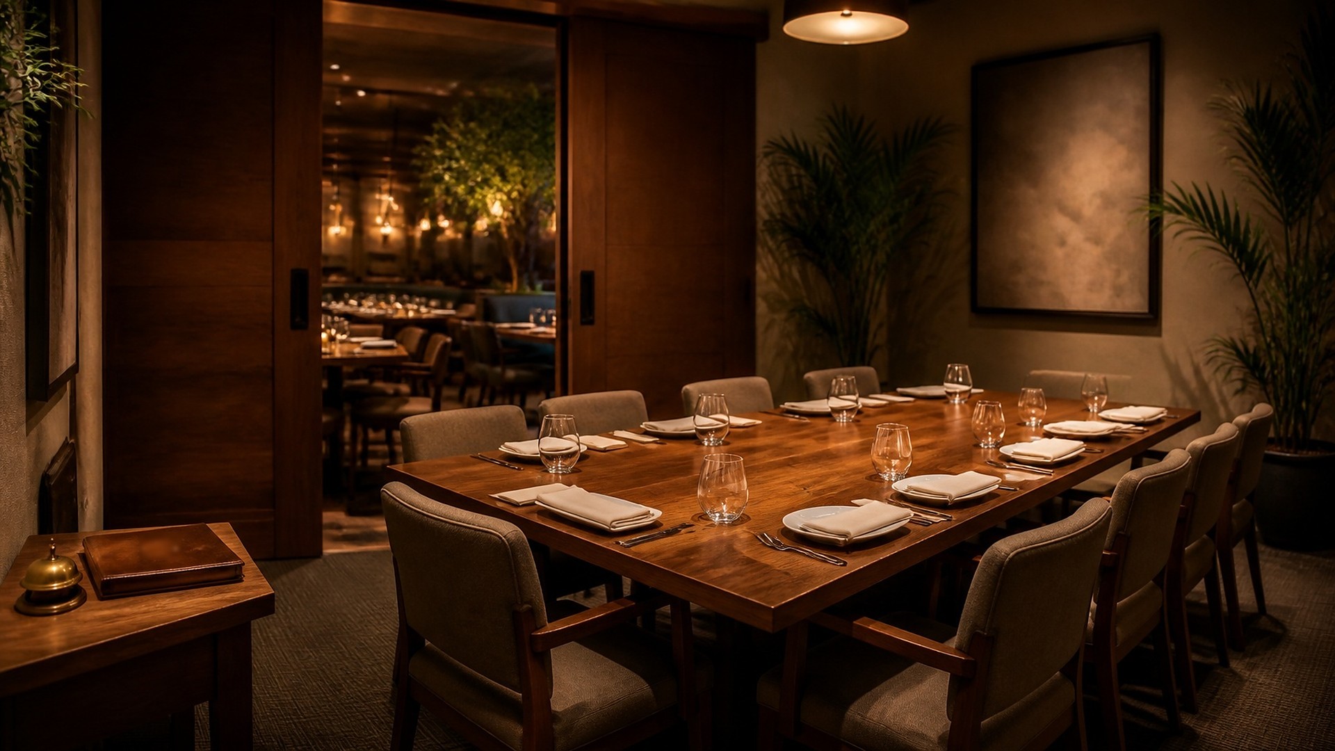 Warm, dark wood private dining room set for a group: a long table with white plates, folded napkins, and glasses, upholstered chairs, potted palms, and a blank framed artwork; open wooden sliding doors show a softly blurred main dining room with pendant lights and a tree; on a side table, a closed leather book and a brass service bell; no people, no logos, no readable text